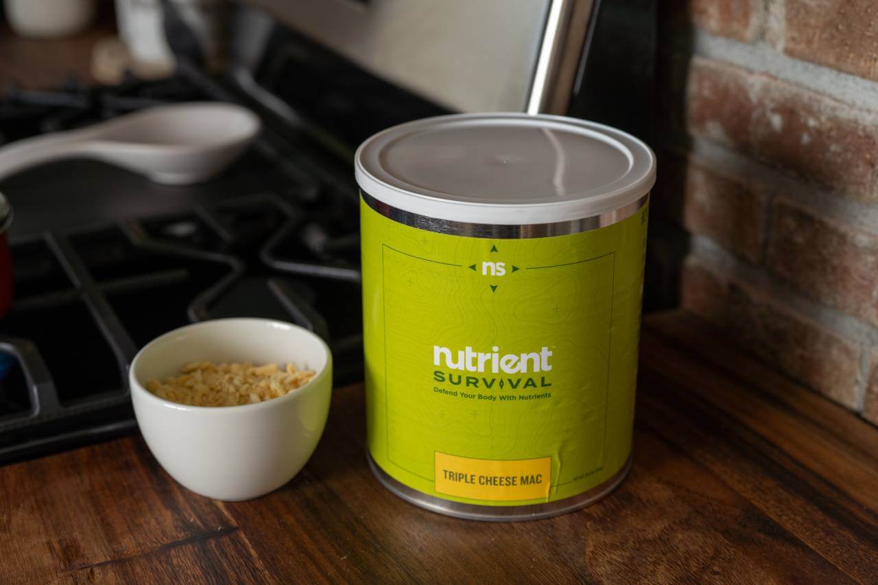 A can of Nutrient Survival Triple Cheese Mac 10 Servings - (SHIPS IN 2-4 WEEKS) is sitting next to a cup of coffee.