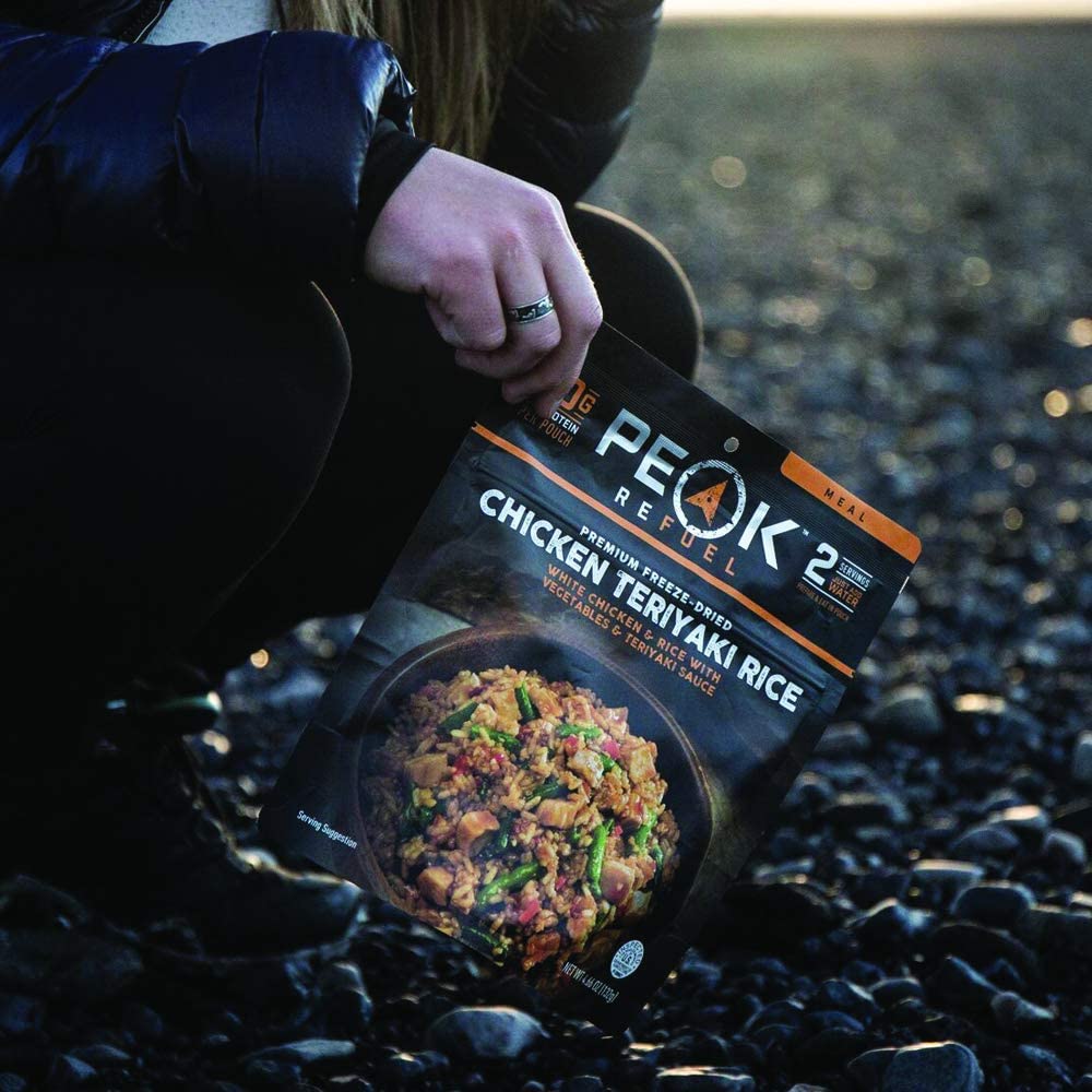 A woman is kneeling on a rocky beach with a Peak Refuel Freeze-Dried Entree Variety Pack Food Storage and Backpacking Food Kit - (SHIPS IN 1-2 WEEKS) of chicken teriyaki rice.