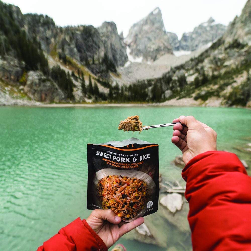 A person holding a Peak Refuel Freeze-Dried Entree Variety Pack Food Storage and Backpacking Food Kit - (SHIPS IN 1-2 WEEKS) in front of a lake.