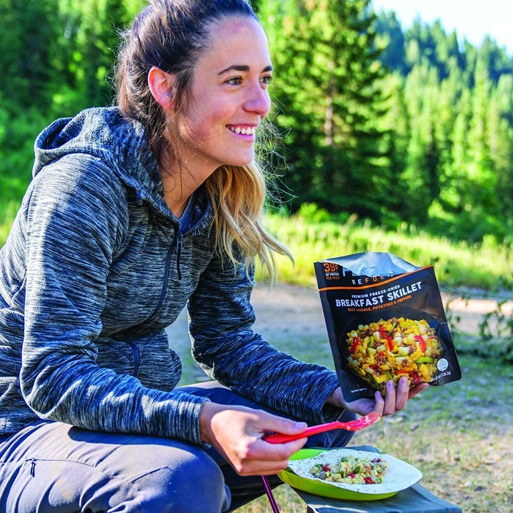 A woman sits on a bench and eats a Peak Refuel Traverse Pack Variety Meals Food Storage and Backpacking Food Kit (SHIPS IN 1-2 WEEKS).