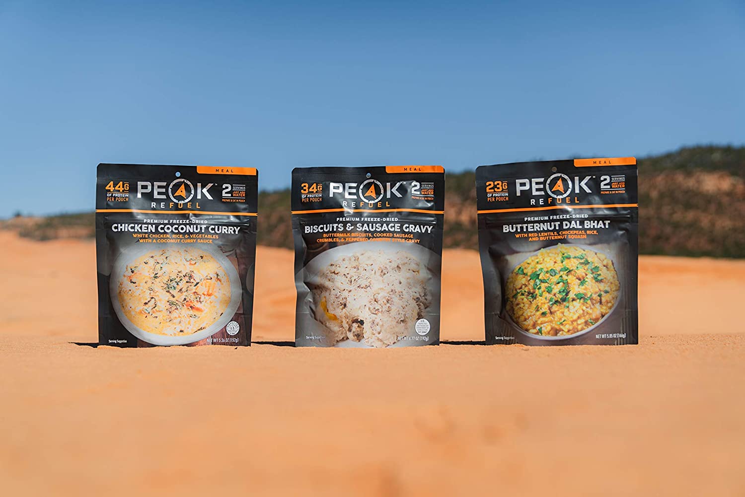 Three packages of Peak Refuel Freeze-Dried Breakfast, Lunch, and Dinner Sampler Food Storage and Backpacking Food Kit - (SHIPS IN 1-2 WEEKS) sitting on top of a sand dune.