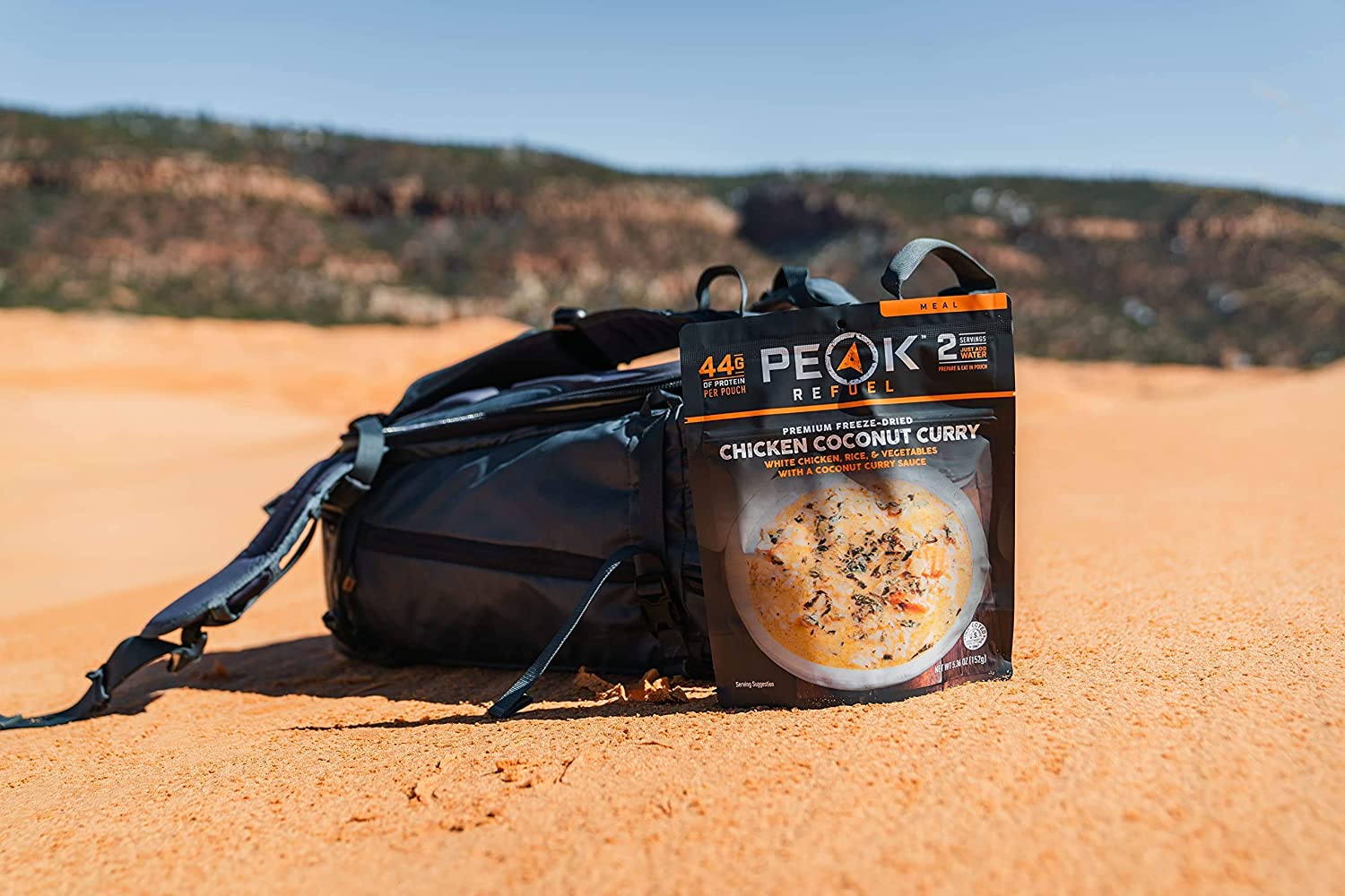 A backpack with a Peak Refuel Freeze-Dried Breakfast, Lunch, and Dinner Sampler Food Storage and Backpacking Food Kit - (SHIPS IN 1-2 WEEKS) sitting on top of a sand dune.