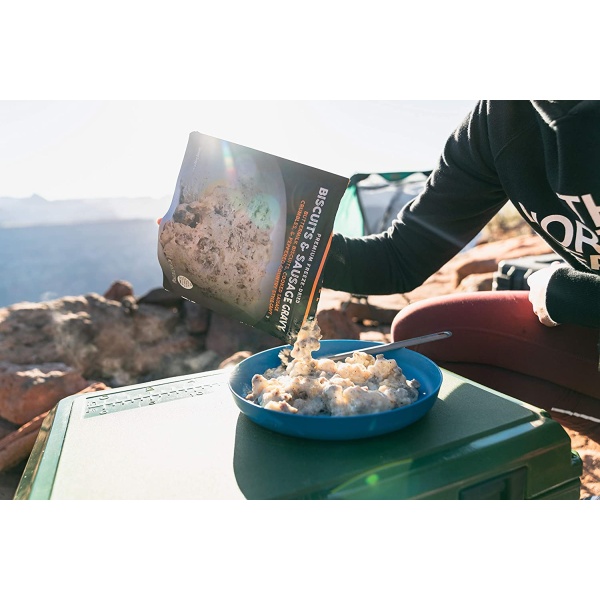 A person eating a bowl of Peak Refuel Freeze-Dried Breakfast, Lunch, and Dinner Sampler Food Storage and Backpacking Food Kit - (SHIPS IN 1-2 WEEKS) on top of a mountain.
