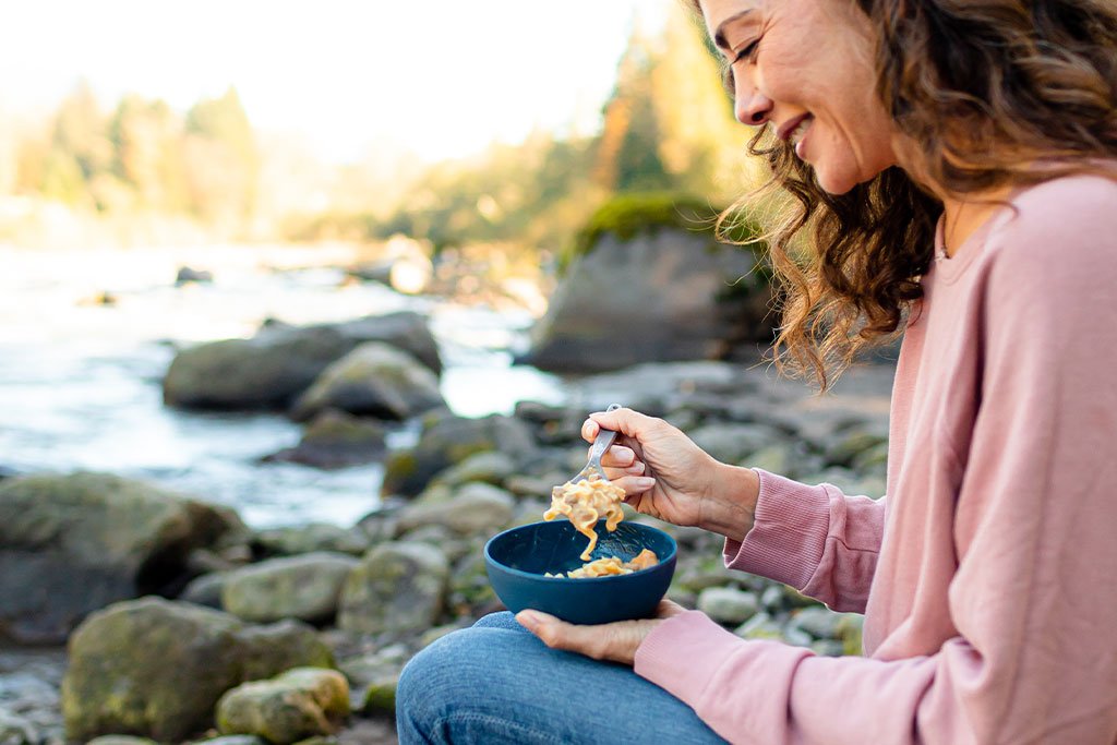 A woman eating a Mountain House Beef Stroganoff Mylar Pouch - 2 Servings - (SHIPS IN 1-2 WEEKS) near a river.