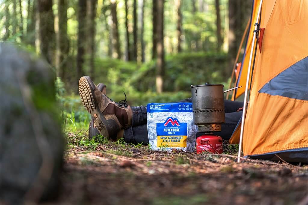 A person sits in front of a Mountain House Gluten-Free Spicy Southwest Breakfast Hash Mylar Pouch - 2 Servings - (SHIPS IN 1-2 WEEKS) in the woods.