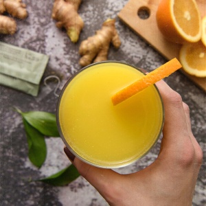 A person holding a glass of orange juice with a slice of ginger, enhanced with Nutrient Survival Immune Boost Drink Mix.