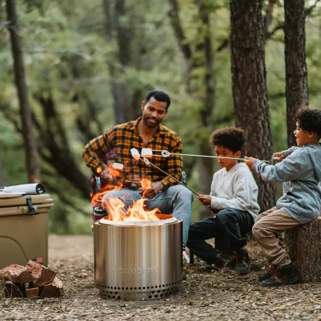 A man and his children are sitting around a portable, stainless steel campfire in the woods.