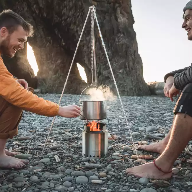 Two men cooking on a portable camping stove on the beach.
