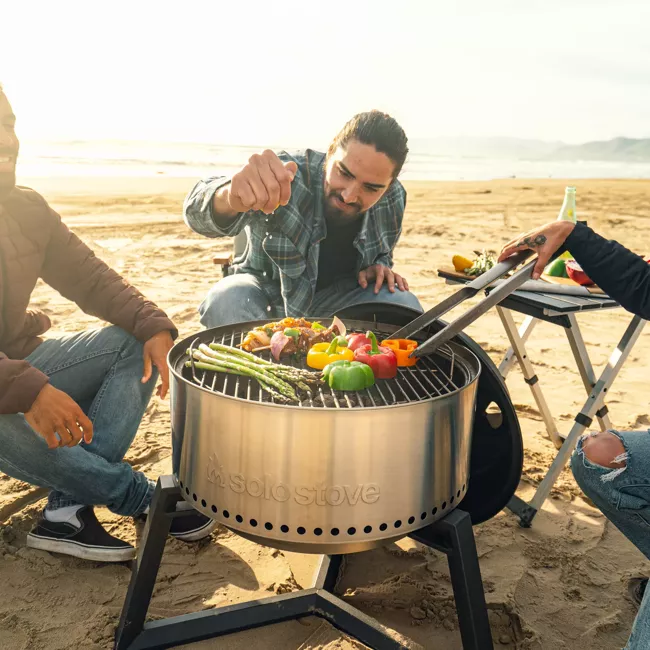 A group of people sitting around a portable grill on the beach.