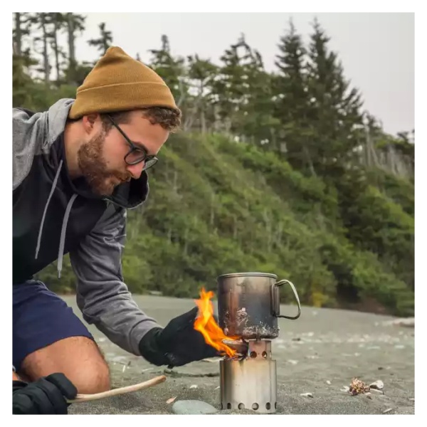 A man is using a portable stainless steel camp stove to cook on an open fire on the beach.