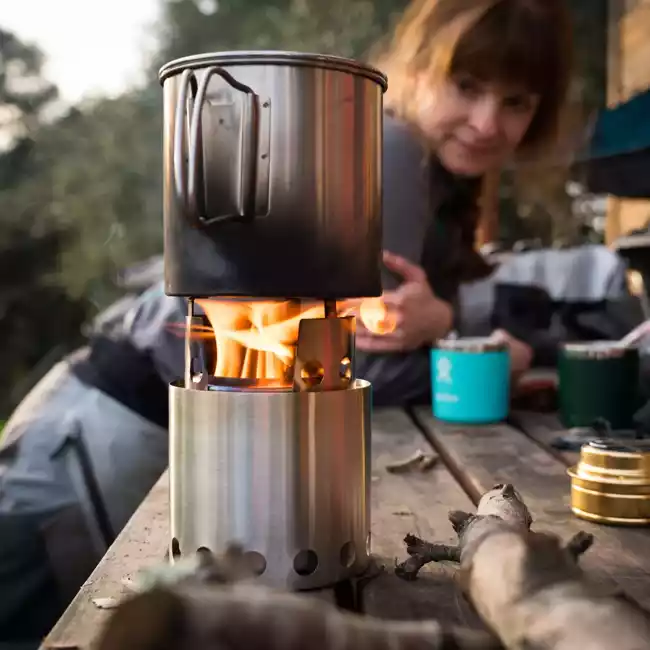 A woman is sitting next to a portable camp stove on a wooden table.