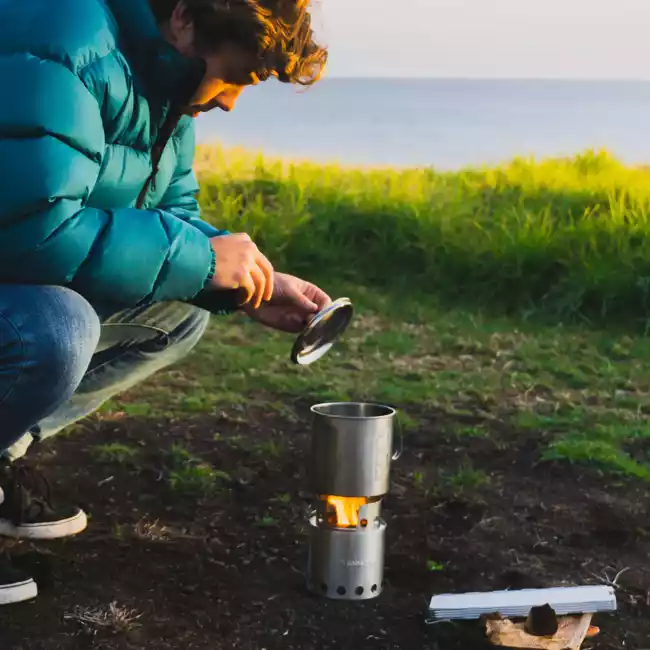A man is cooking on a portable and stainless steel camp stove near the ocean.
