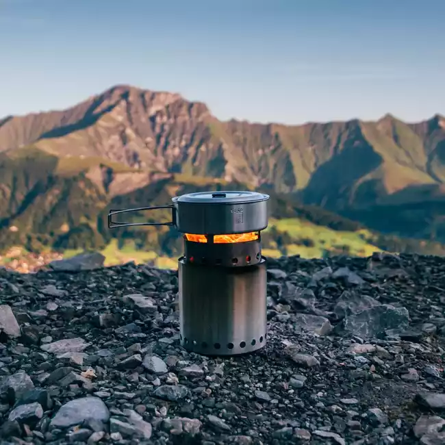 A portable stainless steel stove on top of a mountain, surrounded by mountains in the background.
