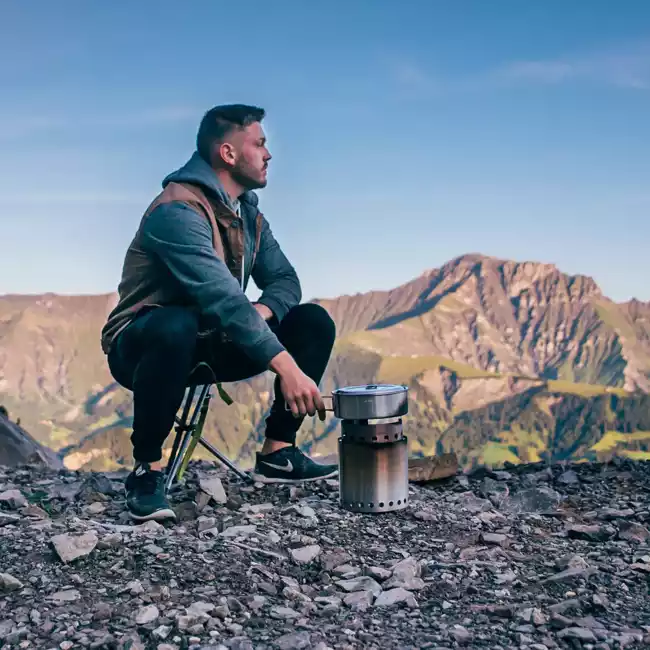 A man sitting on top of a mountain with a portable camp stove.