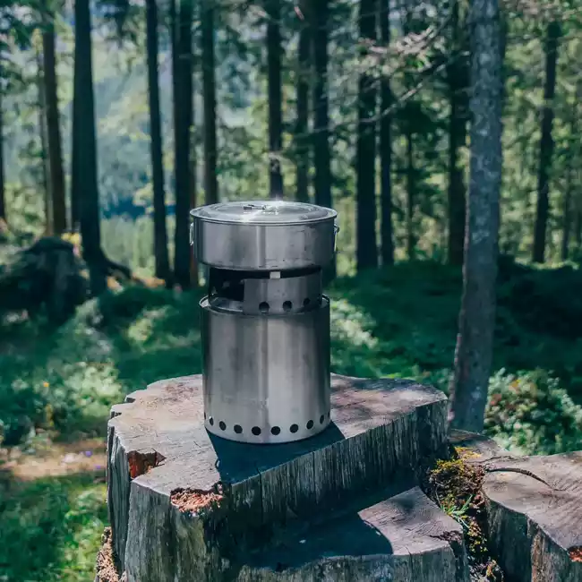 A portable stainless steel camp stove perched on a tree stump in the forest, offering a "smokeless" cooking experience.