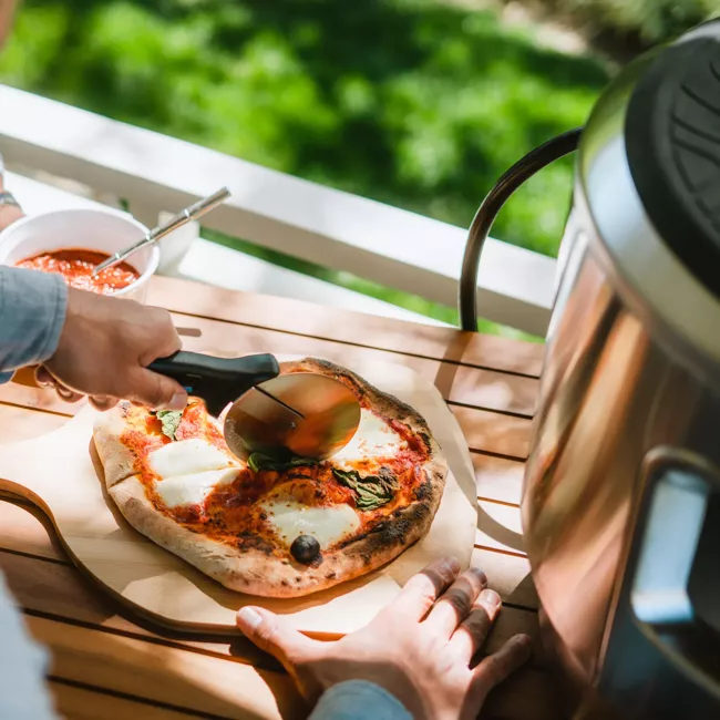 A person is making a pizza on a wooden table using a Solo Stove Pi Essential Bundle Stainless Steel portable grill, known for being "smokeless".
