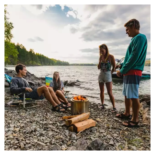 A group of people sitting around a portable and "smokeless" Solo Stove Stainless Steel Ranger Essential Bundle 2.0 on the shore of a lake.