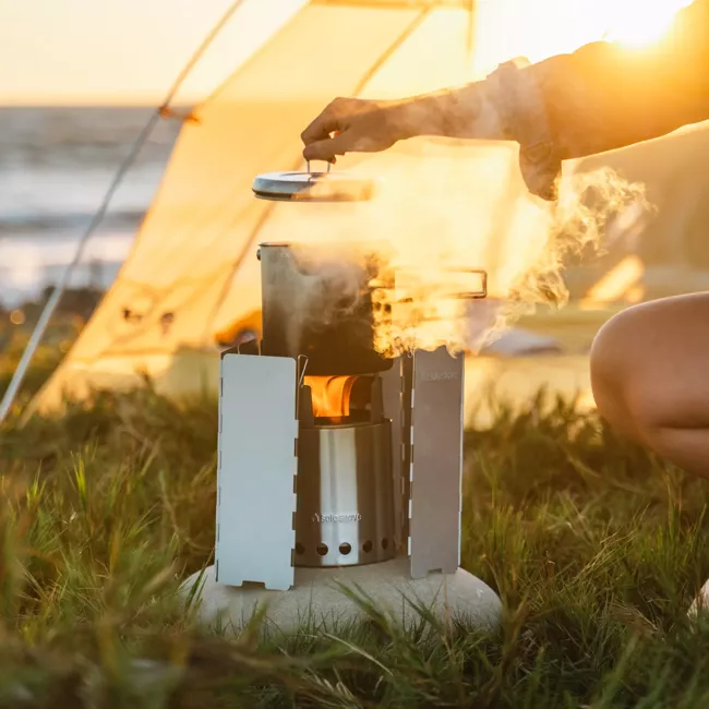 A woman is cooking on a Solo Stove Stainless Steel Titan Camp Stove in front of a tent.