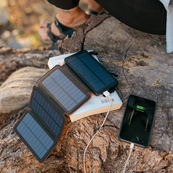 A person sitting on a log with a solar charger ready to charge their phone using the QuadraPro Solar Power Bank from Frog & Co.
