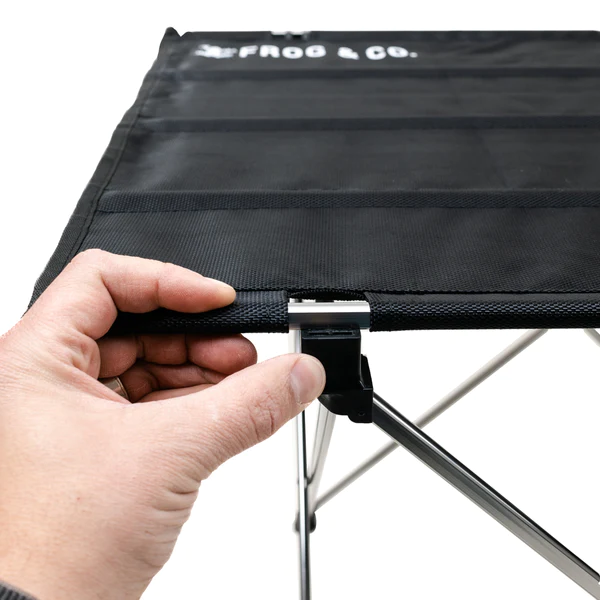 A person is holding a black folding table from Frog & Co.