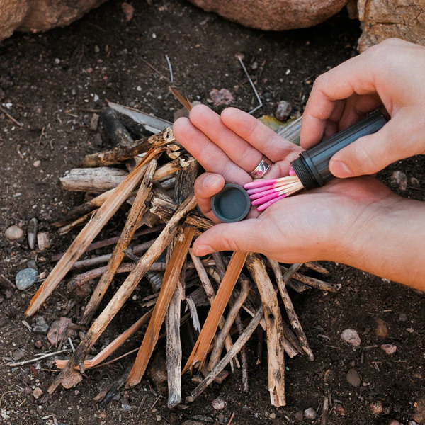 A hand is holding a pink fire starter from Frog & Co in front of a pile of wood, protected by Safety Weatherproof Matches inside a Waterproof Case.