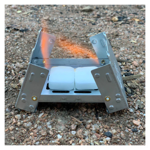 A small camp stove from Frog & Co sitting on the ground, powered by smokeless hexamine fuel tablets.